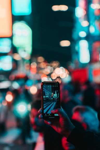 A hand holds a smartphone capturing a vibrant, illuminated cityscape.