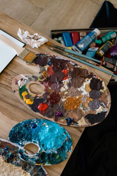 A wooden artist's palette with various paint colours and tools on a table.
