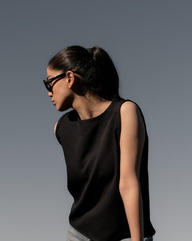Woman in a black sleeveless top and sunglasses, gazing to the side against a grey background.