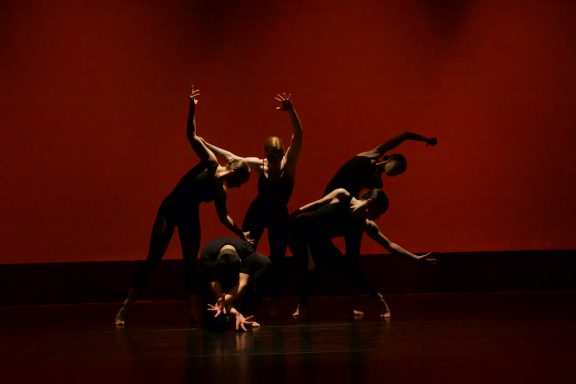 Five dancers in black attire perform expressively against a red backdrop.